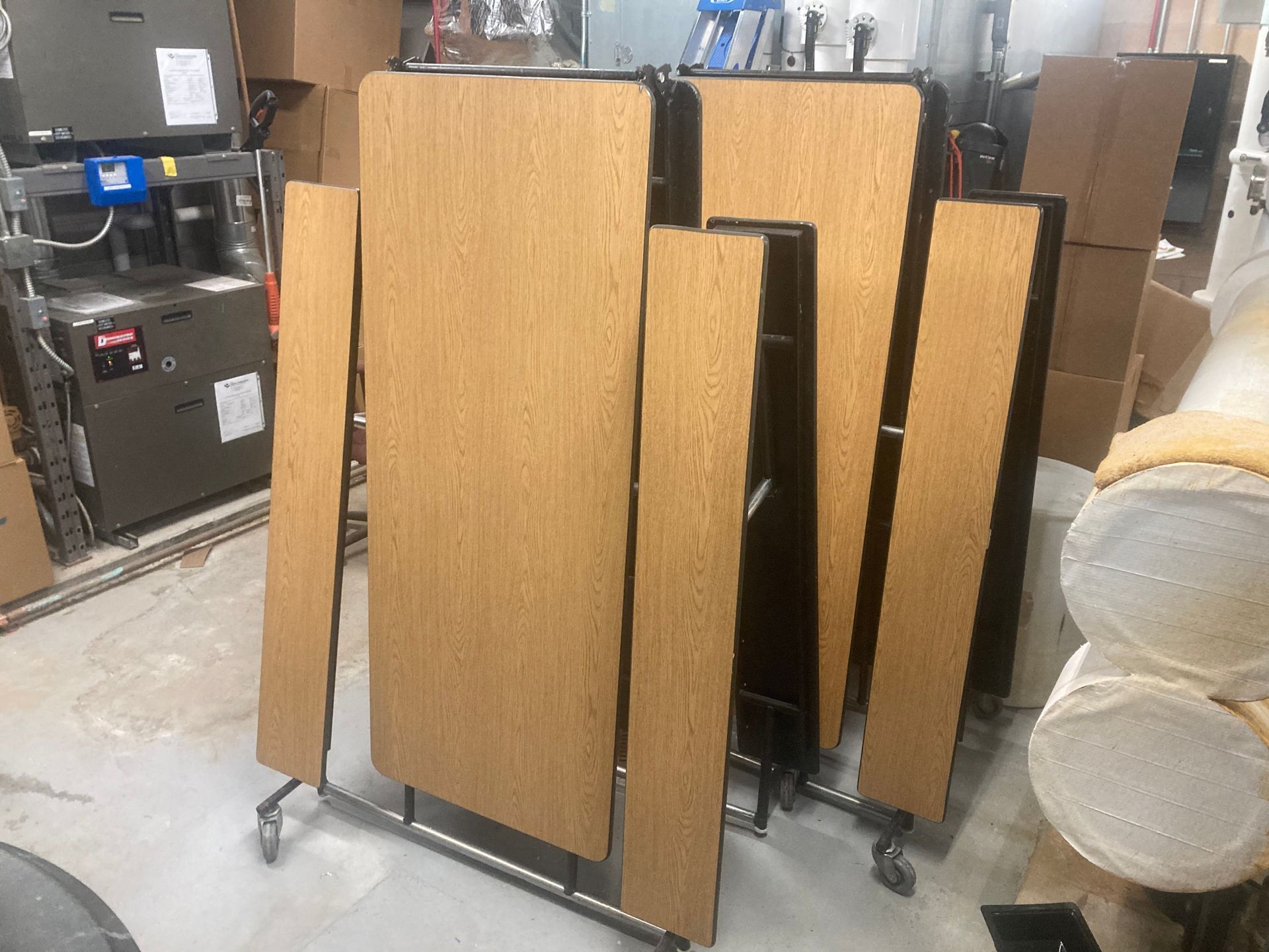 Stack of folding tables with tan tops, black frames, and wheels in a storage room.
