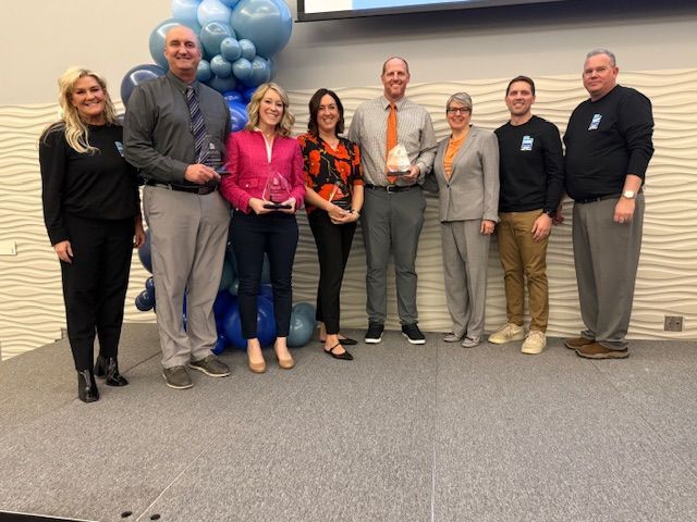 Group of people on stage holding awards; blue and white balloon backdrop.