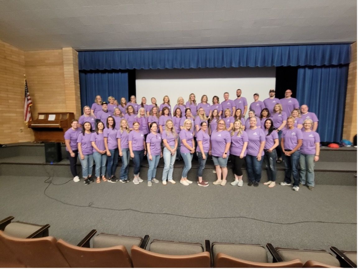 A large group of people wearing purple shirts are standing in front of a stage.