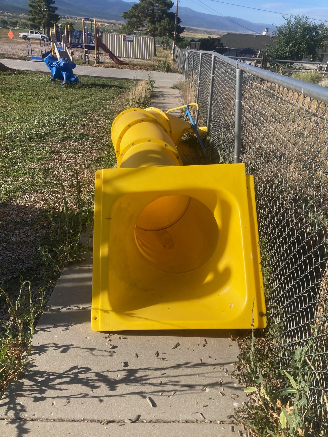 A yellow slide is sitting on the sidewalk next to a chain link fence.
