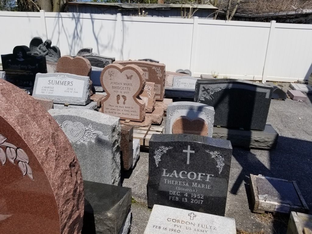 Gravestones of various shapes, sizes, and colors are displayed outdoors in front of a white fence.