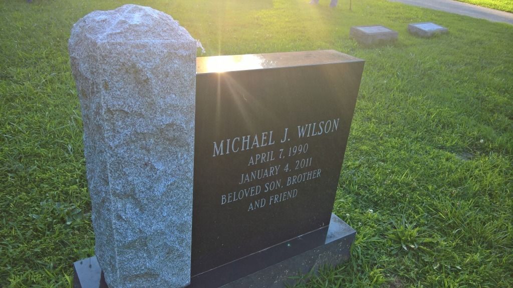 Tombstone with engraved text, next to a rough stone border, in a sunlit grassy cemetery.
