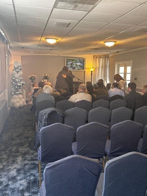A room with rows of chairs facing a person speaking at a podium, possibly a memorial or meeting.