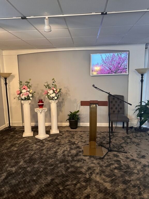 A room set for a service; flowers on pedestals flank a podium, with a TV displaying a floral image.
