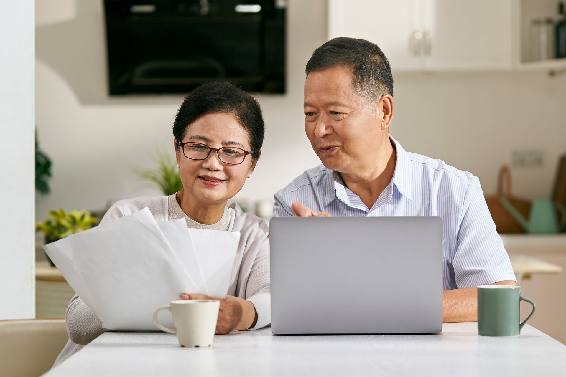 An elderly couple is sitting at a table with a laptop and a notebook.