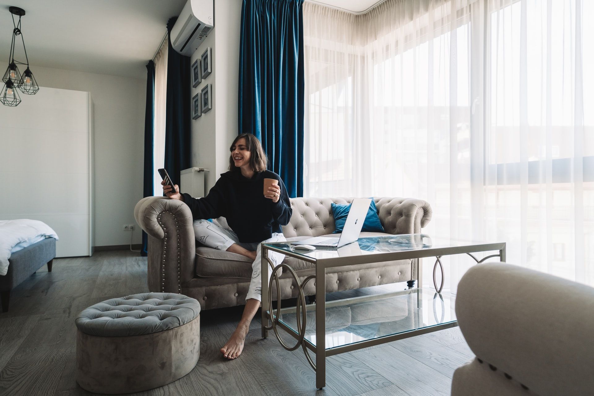 A woman is sitting on a couch in a living room holding a cup of coffee and a remote control.