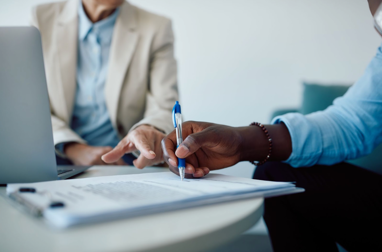 A man and a woman are sitting at a table with a laptop and a pen.