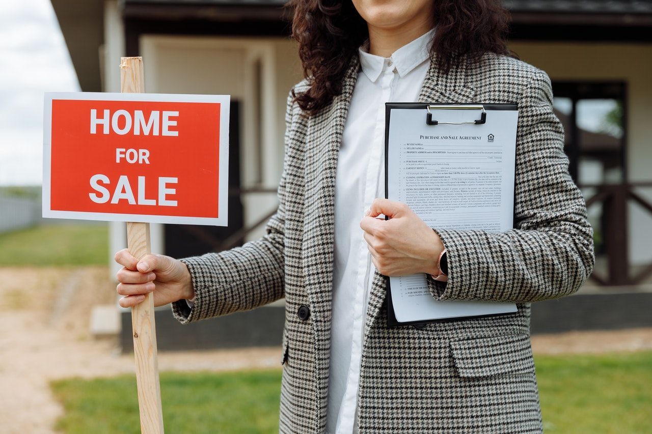 A woman is holding a home for sale sign and a clipboard in front of a house.