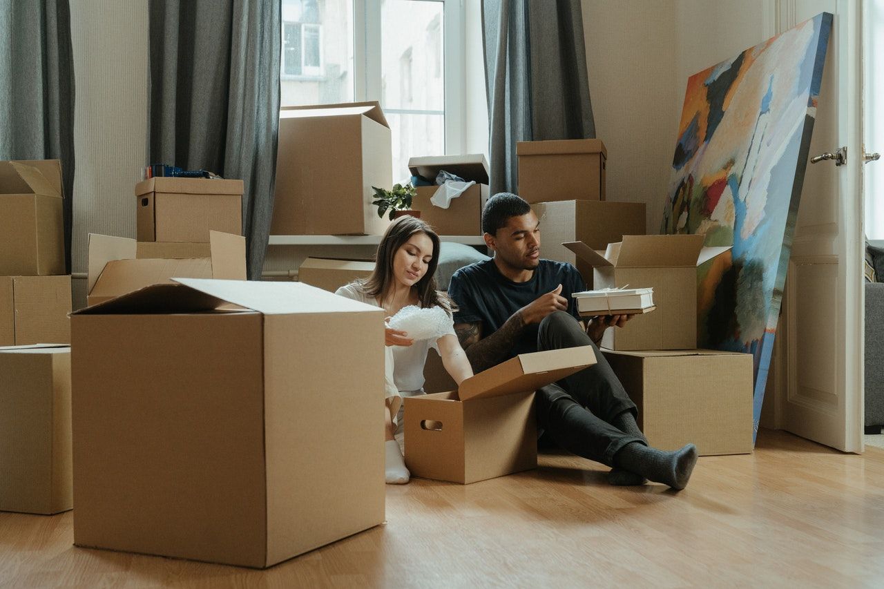 A man and a woman are sitting on the floor in a living room surrounded by cardboard boxes.