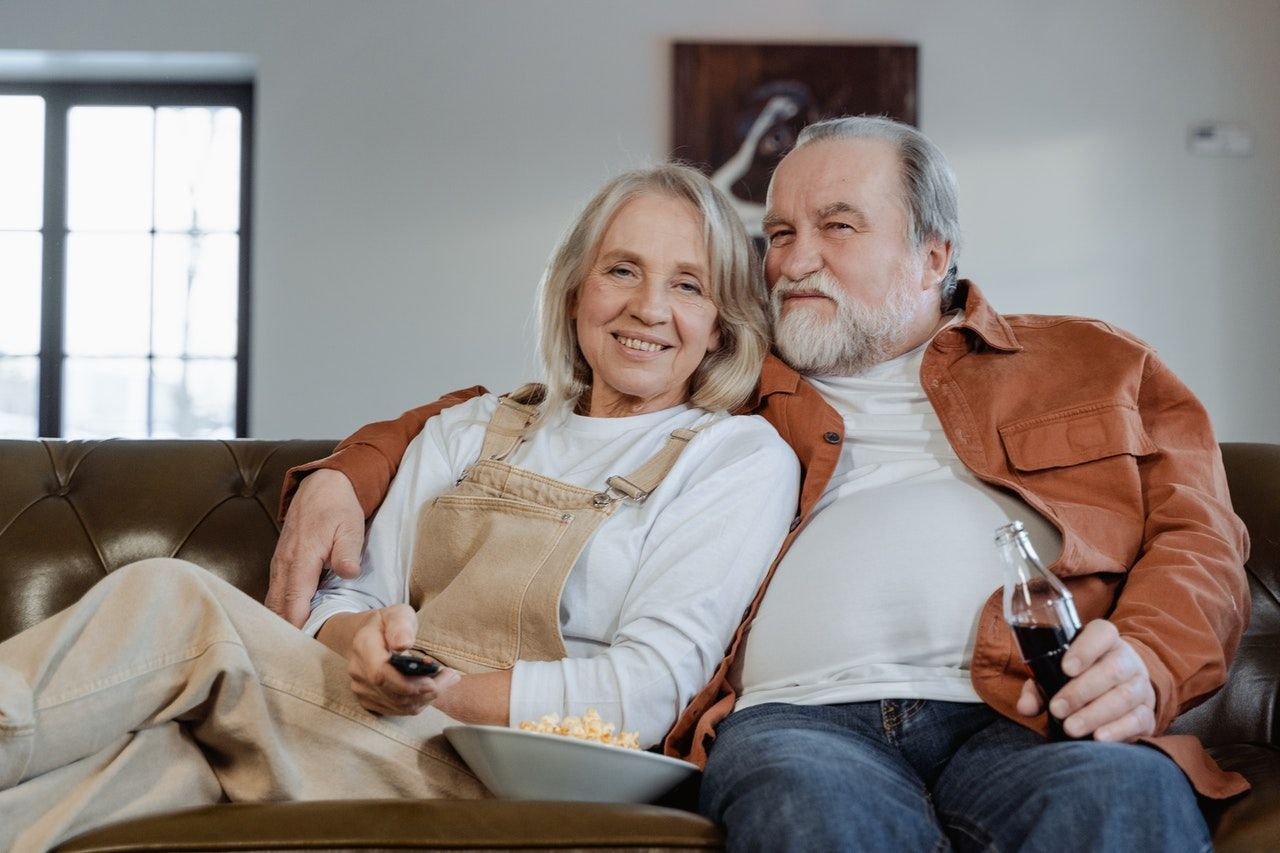 An elderly couple is sitting on a couch watching tv.