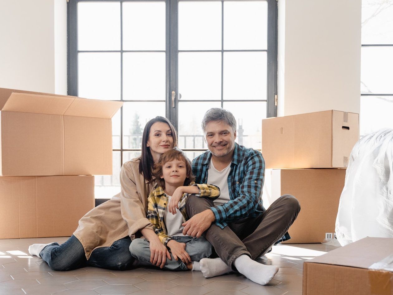 A family is sitting on the floor in their new home surrounded by cardboard boxes.