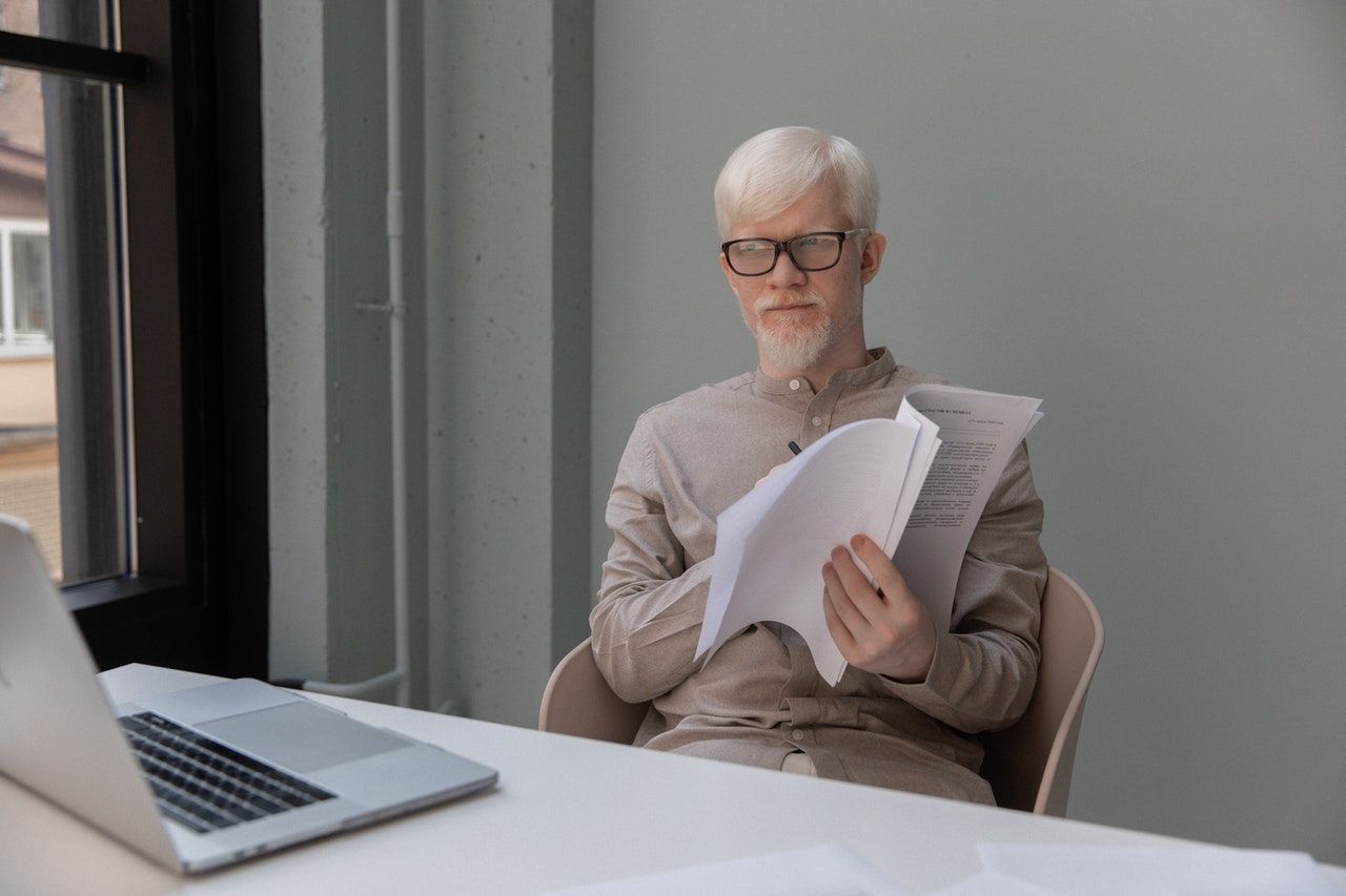 An albino man is sitting at a desk with a laptop and holding a piece of paper.