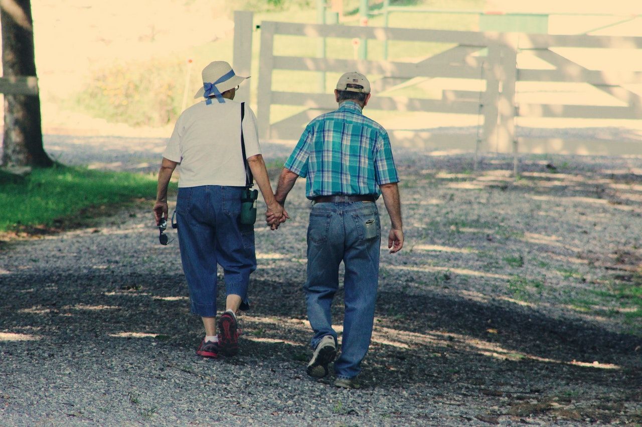 An elderly couple is walking down a dirt road holding hands.