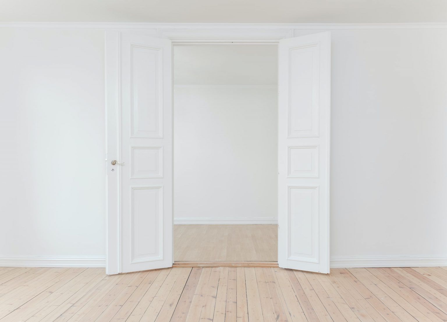 An empty room with white walls and wooden floors and two open doors.