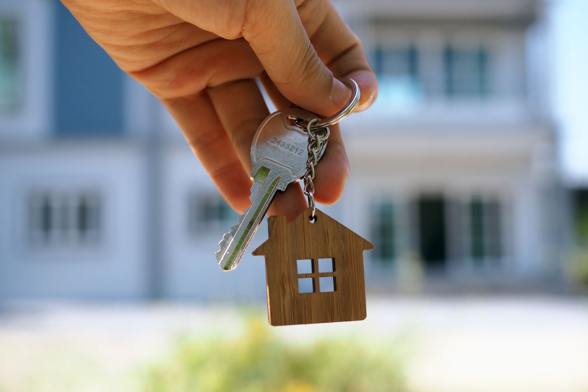 A hand is holding a set of keys with a house keychain in front of a house.