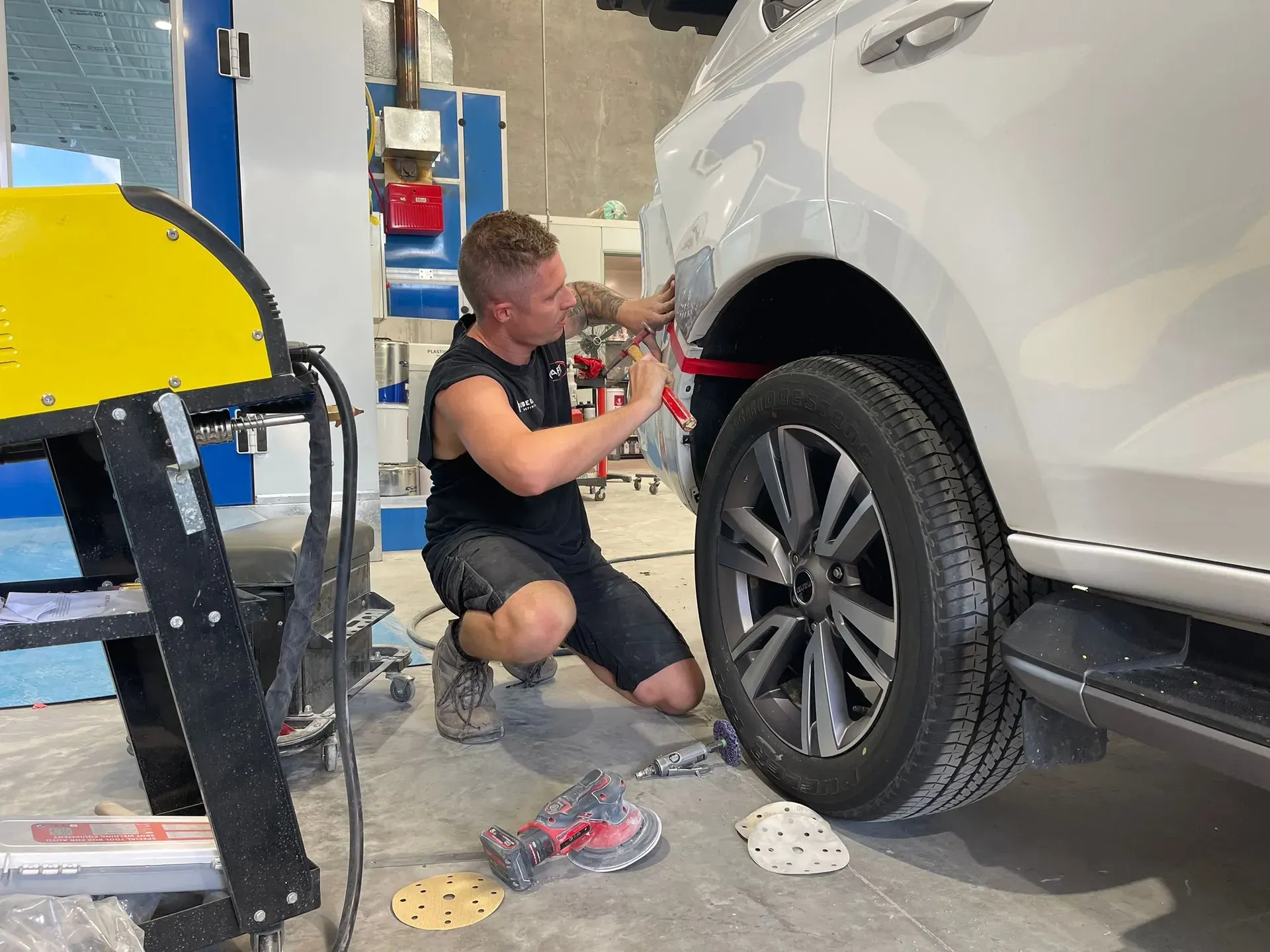 Man repairing a car's rear panel in a workshop. He is using tools and inspecting the damage near the wheel. — Coolum Smash Repairs in Coolum Beach, QLD
