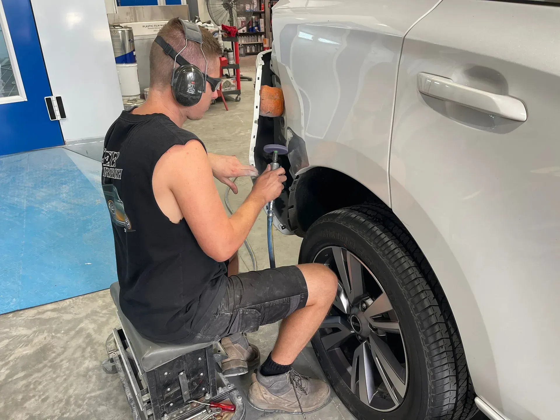 Man repairing car's side panel in a shop; he is seated, wearing ear protection, and holding a tool. — Coolum Smash Repairs in Coolum Beach, QLD