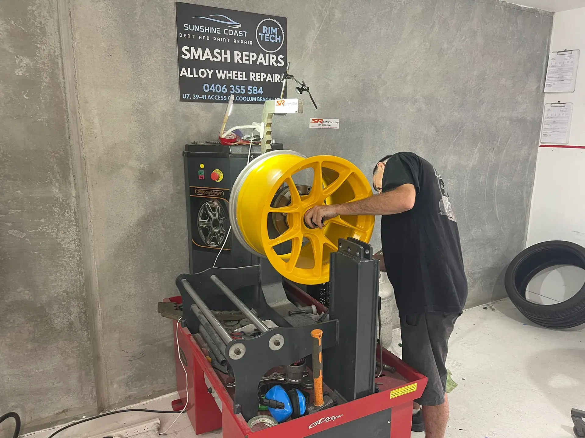 Man balancing a yellow car wheel on a machine in a workshop. Gray walls, tire and tools present. — Coolum Smash Repairs in Coolum Beach, QLD