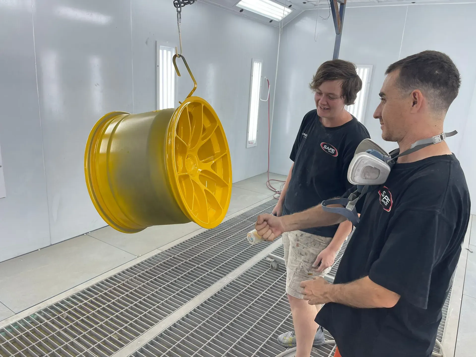 Two men inspecting a yellow car wheel hanging in a paint booth.  — Coolum Smash Repairs in Coolum Beach, QLD