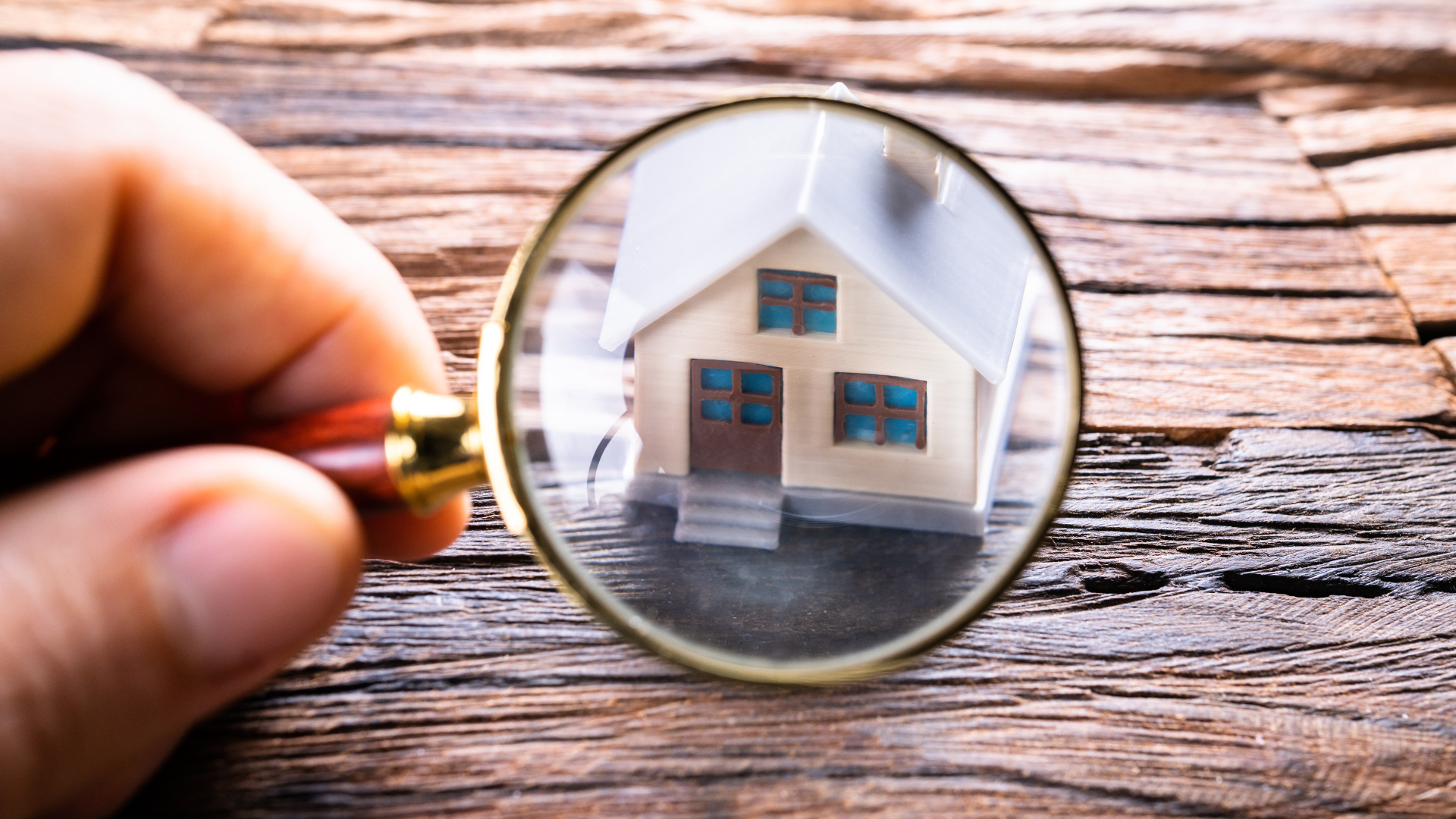 A person is holding a magnifying glass over a model house on a wooden table.