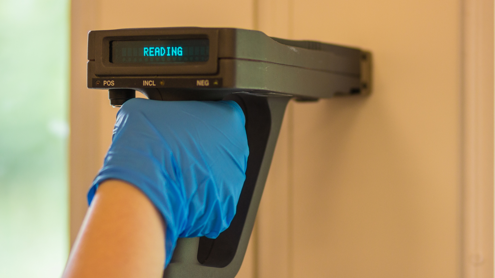 A person wearing blue gloves is holding a device on a wall.