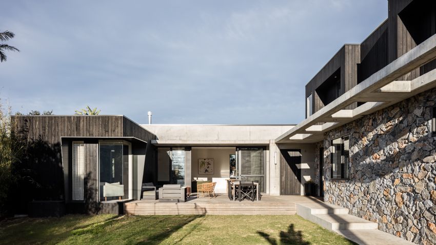 Modern home exterior with stone wall, concrete beams, and dark wood siding, looking onto a patio and green lawn — Make The Mark Building Services in Illawarra, NSW