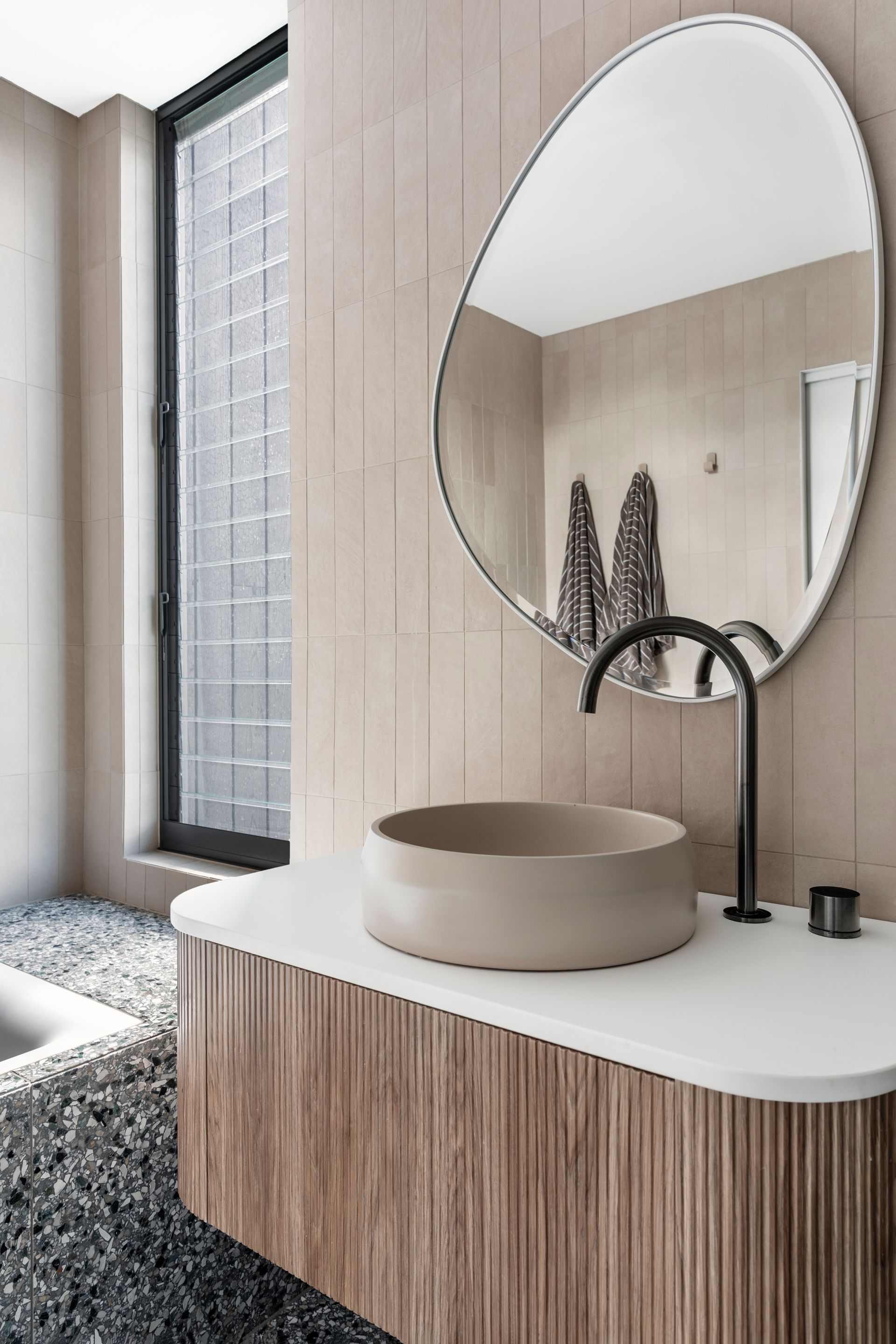 Modern bathroom with round mirror, beige vessel sink, ribbed wooden cabinet, and pebble floor — Make The Mark Building Services in Illawarra, NSW