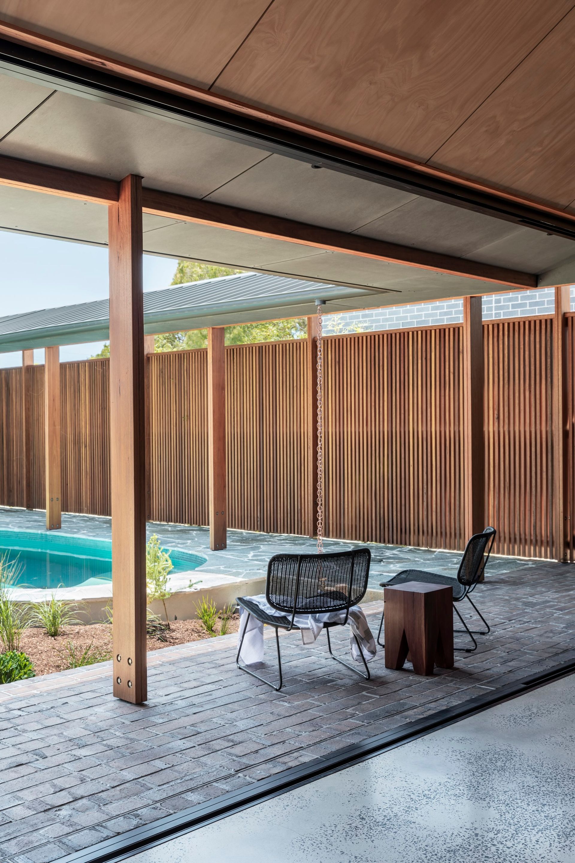 Patio with pool view: wooden posts, gray floor, two chairs, and a small wooden side table — Make The Mark Building Services in Illawarra, NSW