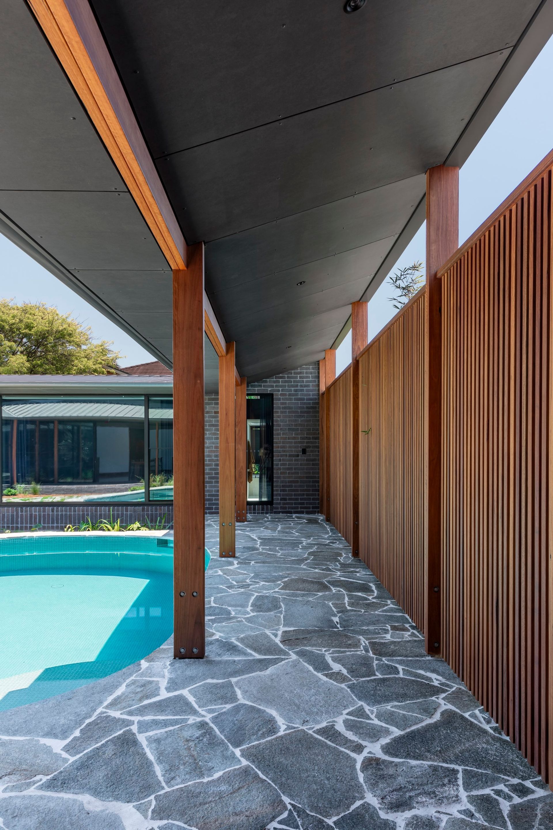 Covered walkway with pool view. Brown columns, wooden wall, gray stone path, and dark ceiling — Make The Mark Building Services in Illawarra, NSW