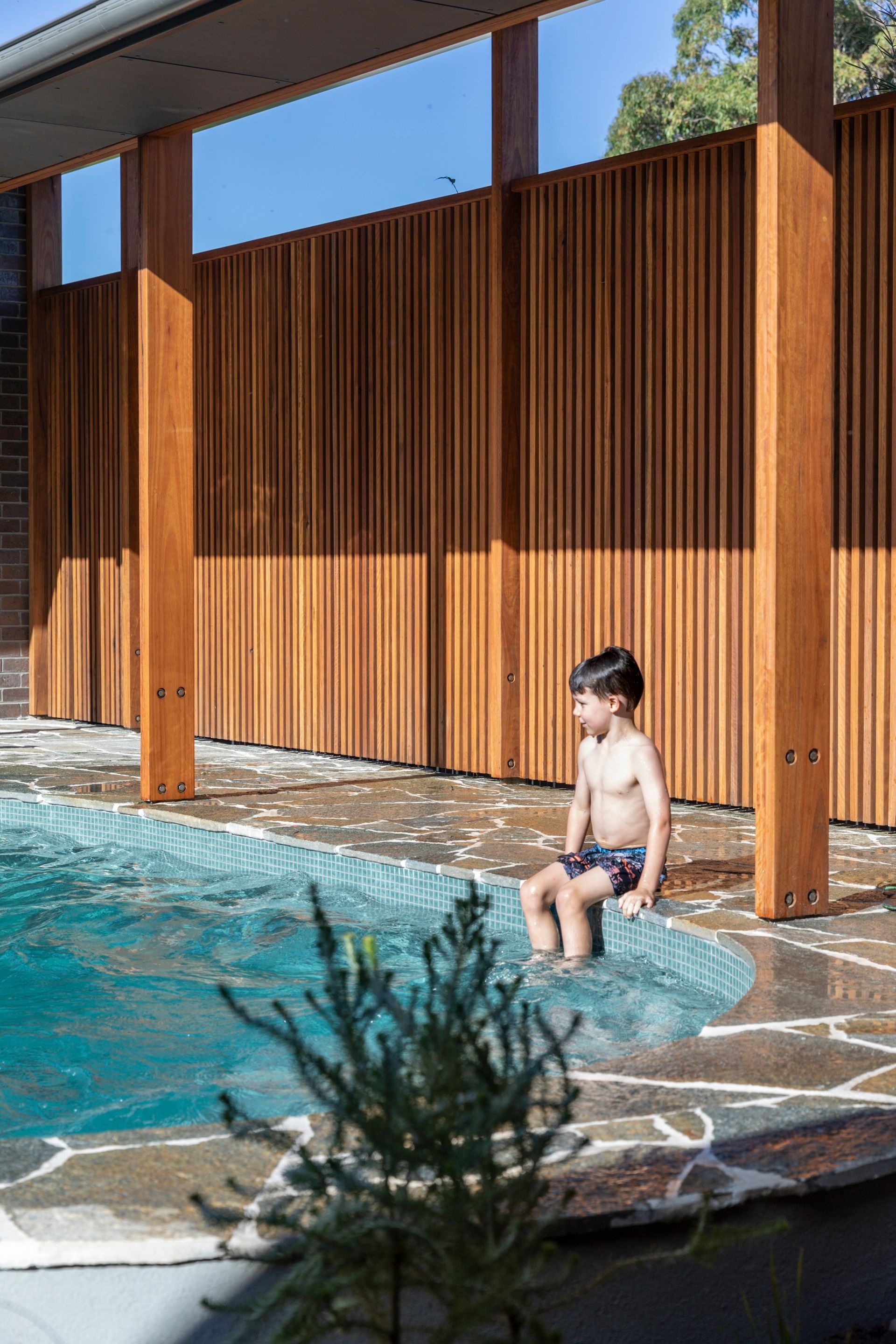 Boy sits on pool edge, feet in water. Wooden fence and posts in background. Sunny day — Make The Mark Building Services in Illawarra, NSW