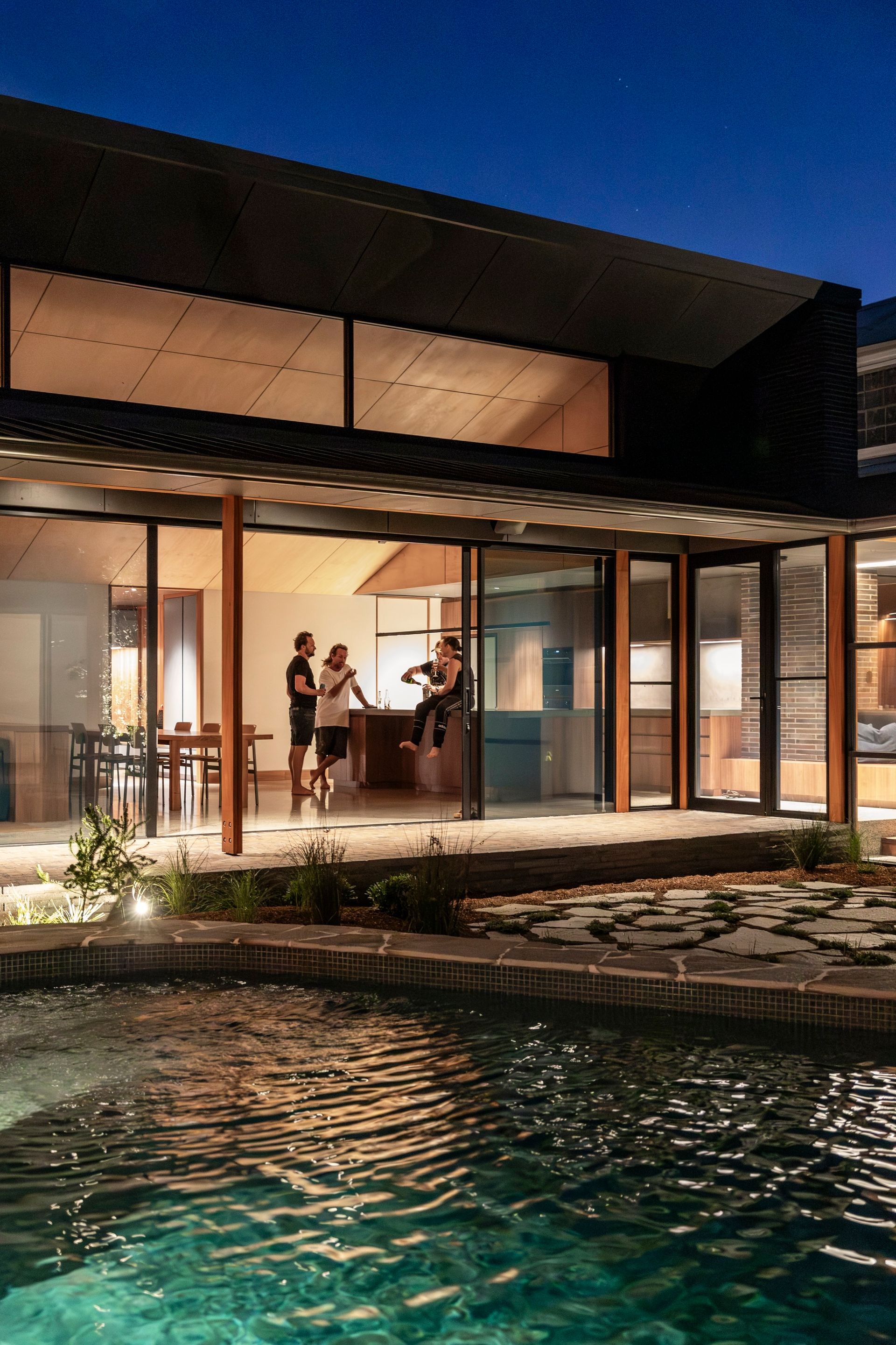 Night scene: People inside a modern house with open glass walls. Pool in the foreground — Make The Mark Building Services in Illawarra, NSW