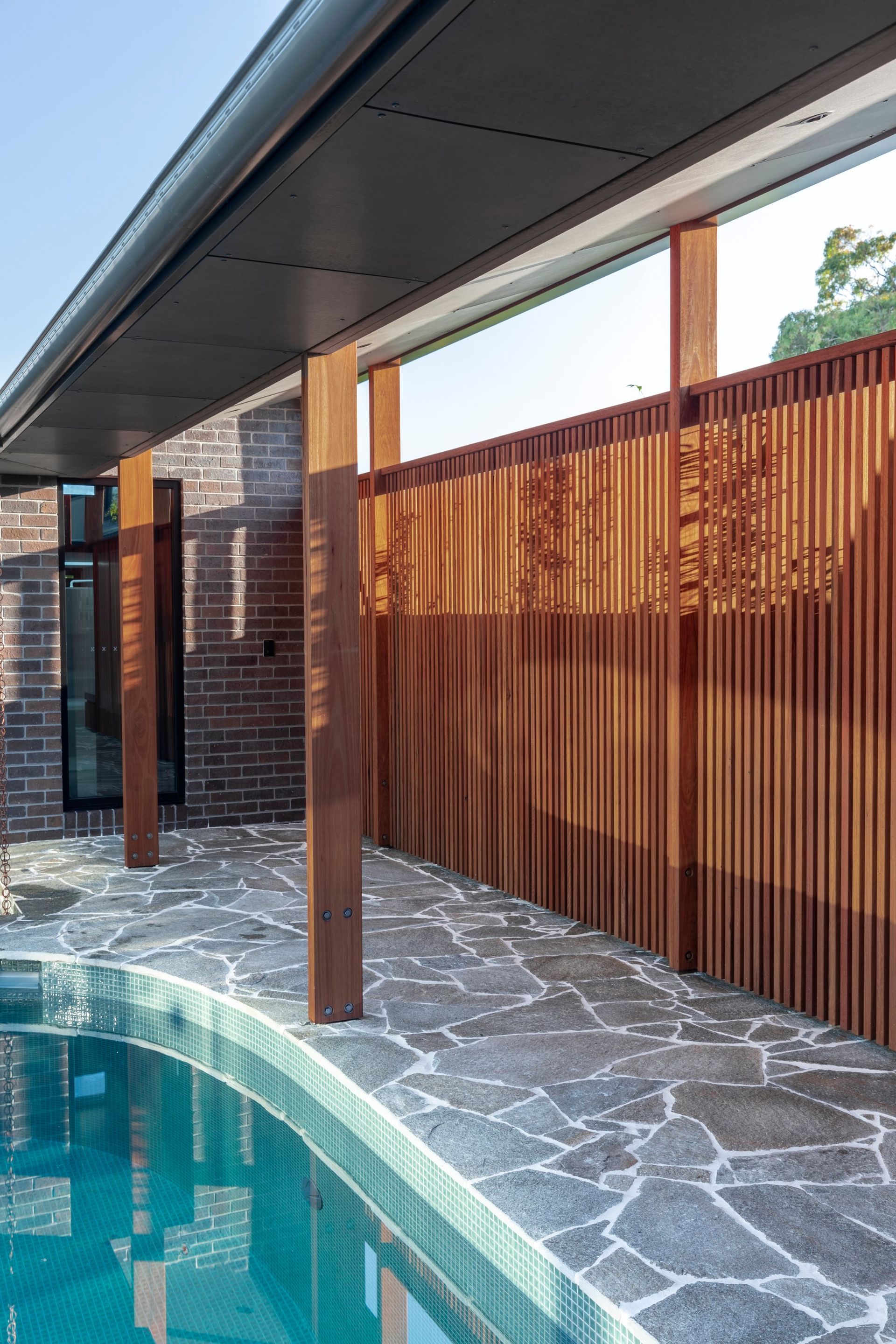 Poolside view with wooden pillars, a vertical slat fence, and a dark stone walkway. Blue pool water — Make The Mark Building Services in Illawarra, NSW