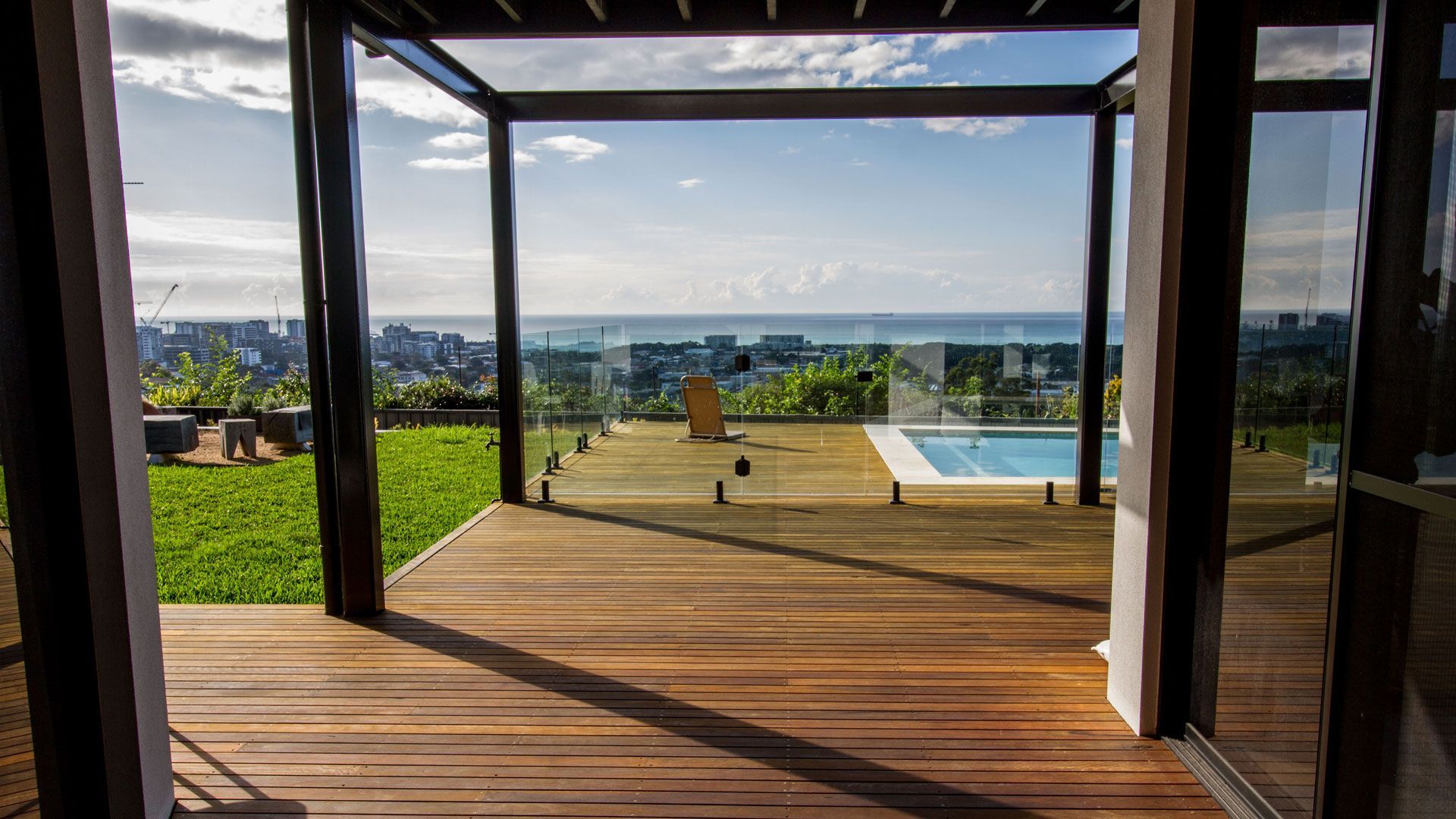Wooden deck overlooking a city, ocean, and pool framed by a pergola and glass walls. Sunny day  — Make The Mark Building Services in Illawarra, NSW