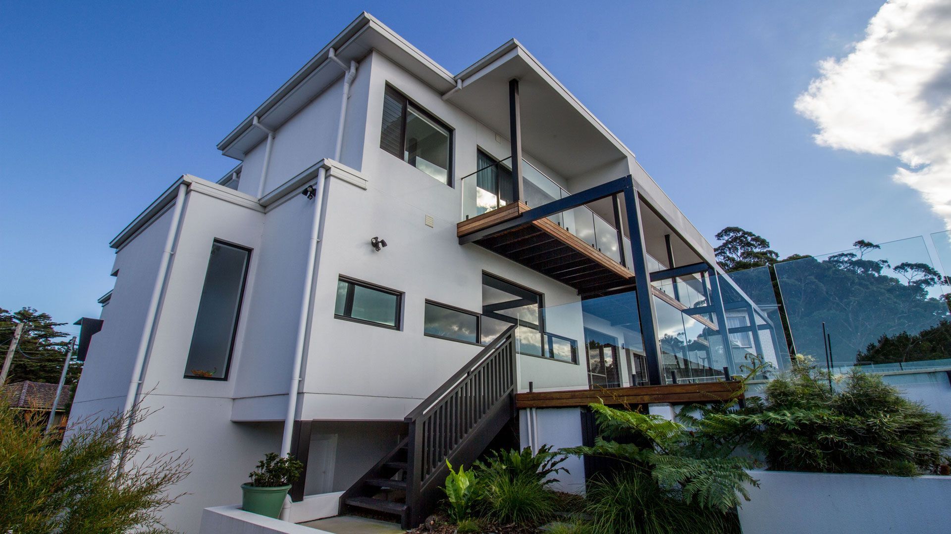 Modern white house with dark wood deck and stairs, glass railings, against a blue sky  — Make The Mark Building Services in Illawarra, NSW