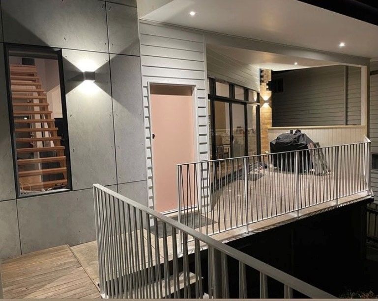 Exterior view of a deck with railing, a light pink door, and a staircase. Two wall sconces illuminate the scene - Make The Mark Building Services in Mangerton, NSW