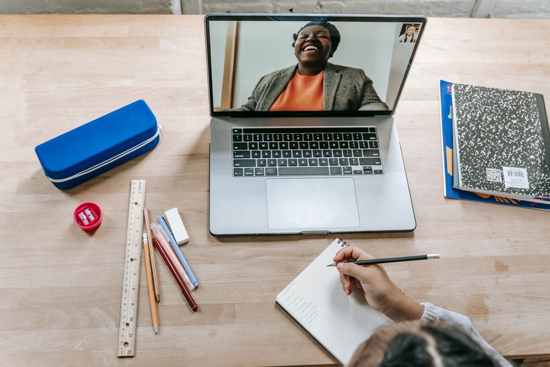 Laptop on desk with video call, student taking notes. Blue pencil case, pencils, ruler, and notebooks visible.