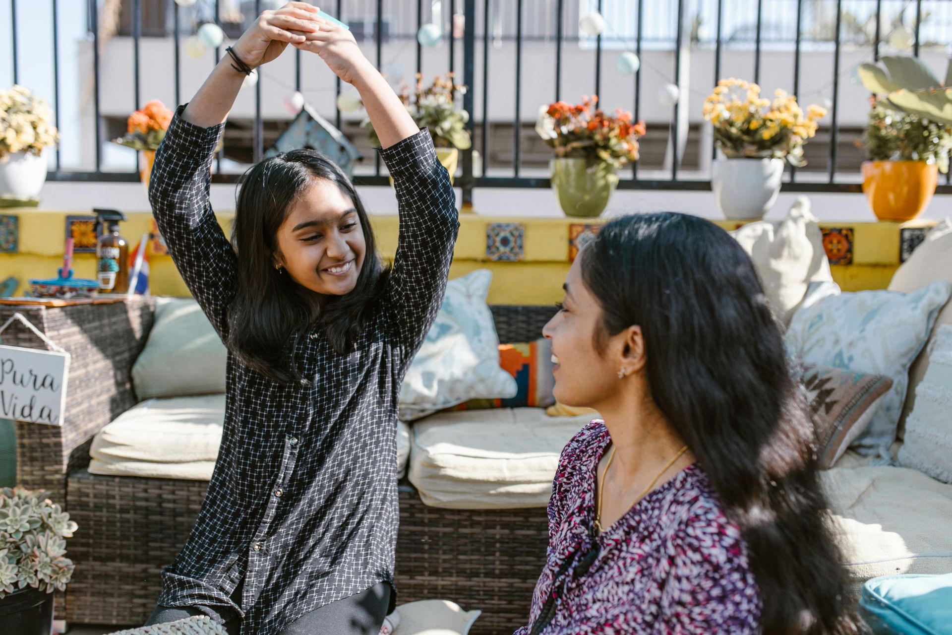 Two women on a patio. One raises her arms, smiling. The other looks on, also smiling. Plants in pots are visible.