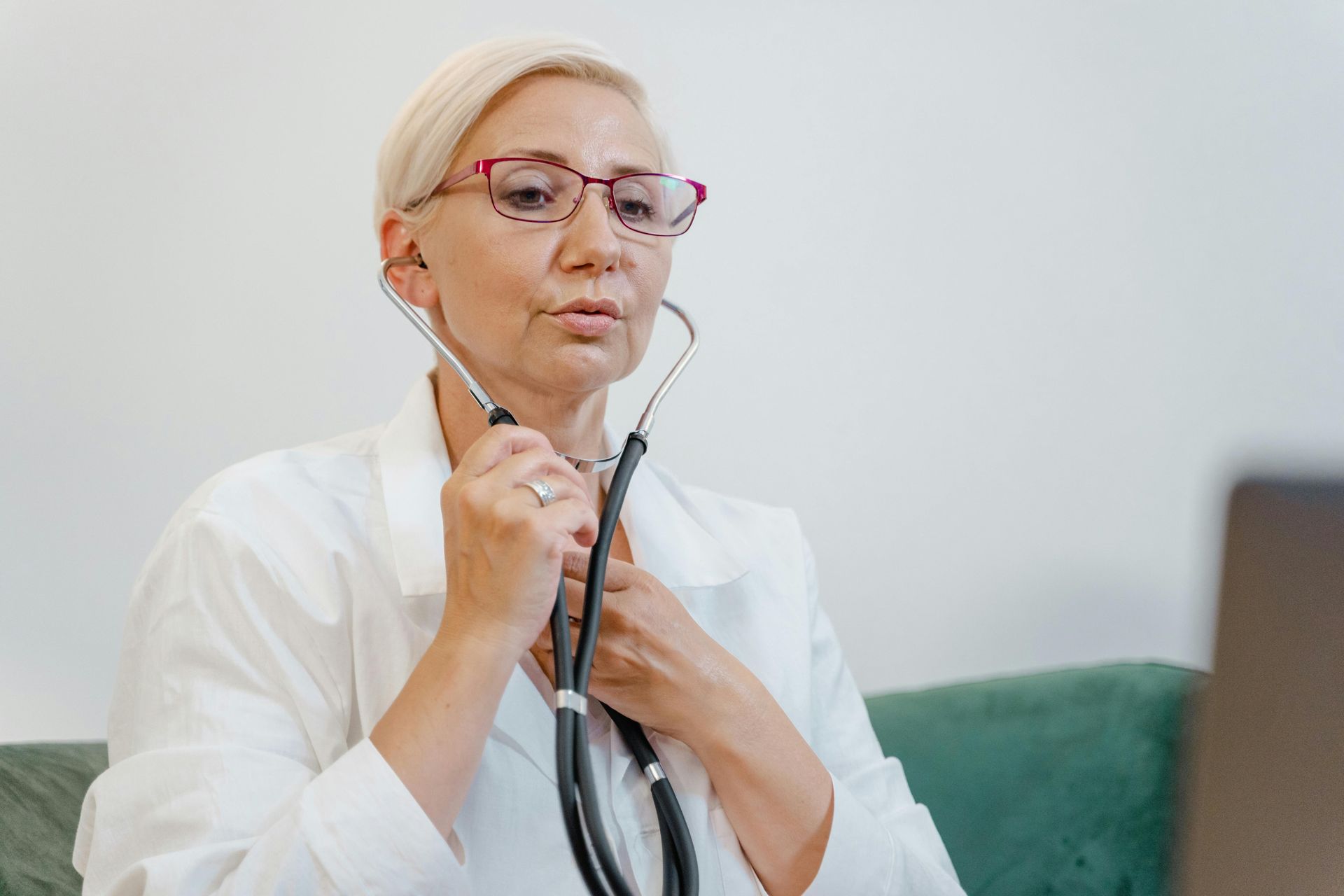 Doctor with stethoscope using laptop. Blonde hair, glasses, white coat, teal sofa.