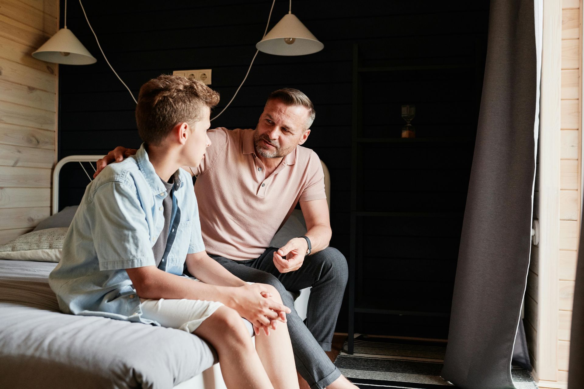 Man comforting teen in bedroom; man's hand on teen's shoulder.