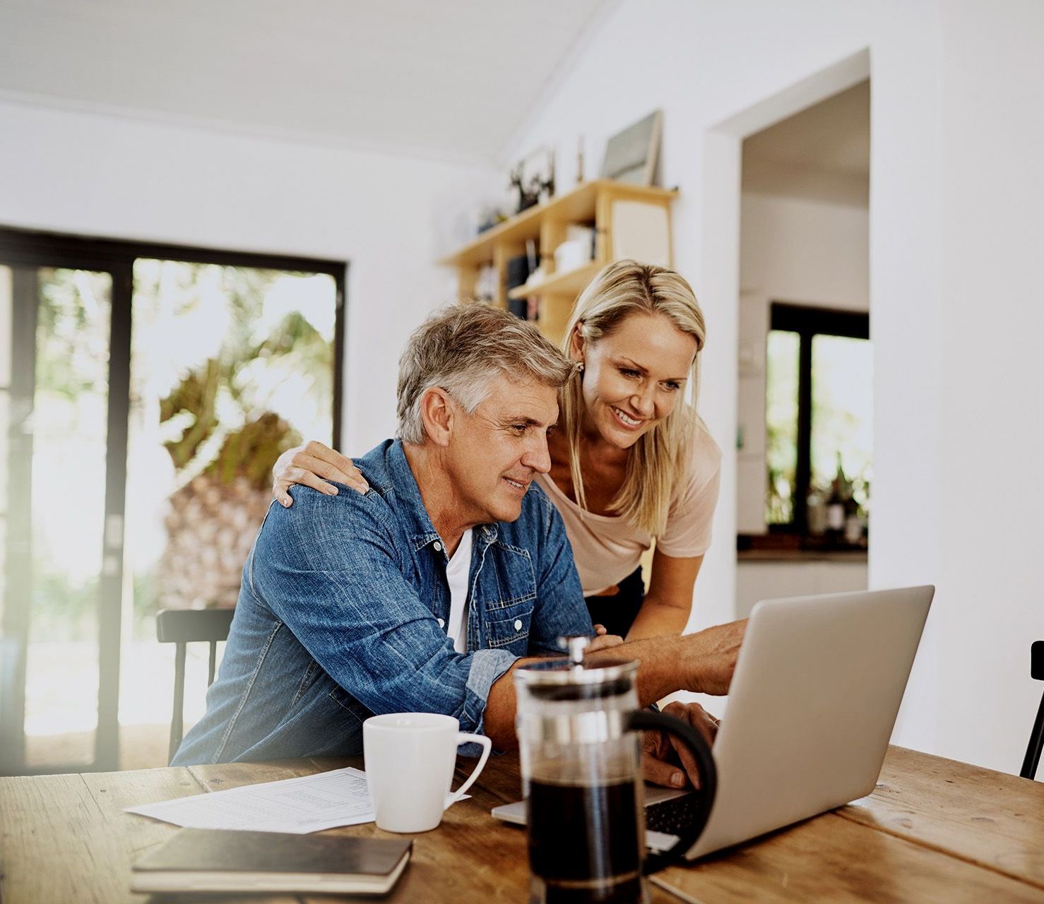 A couple smiling while looking at a laptop together at a wooden dining table, with a coffee press and mug nearby.