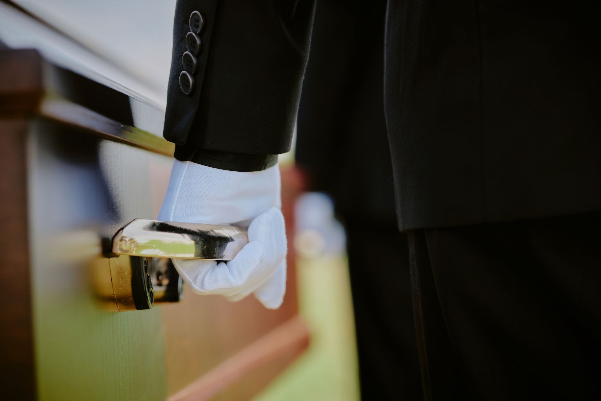 A person in a black suit and white glove holds the handle of a wooden casket.