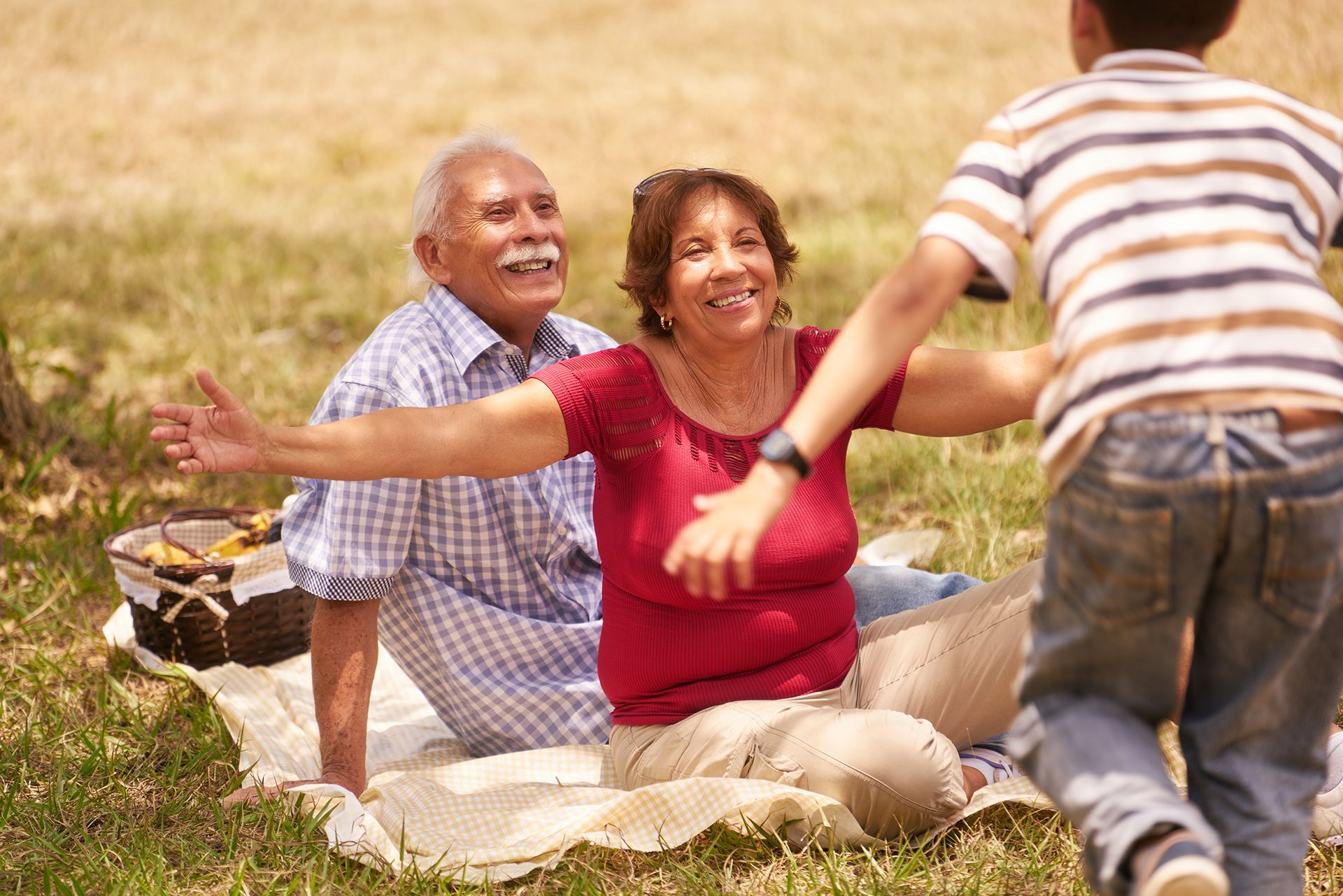 An older couple sitting on a picnic blanket with arms open to embrace a child running toward them.