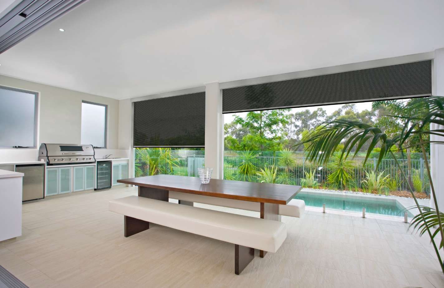 A dining room with a table and benches and a swimming pool in the background.