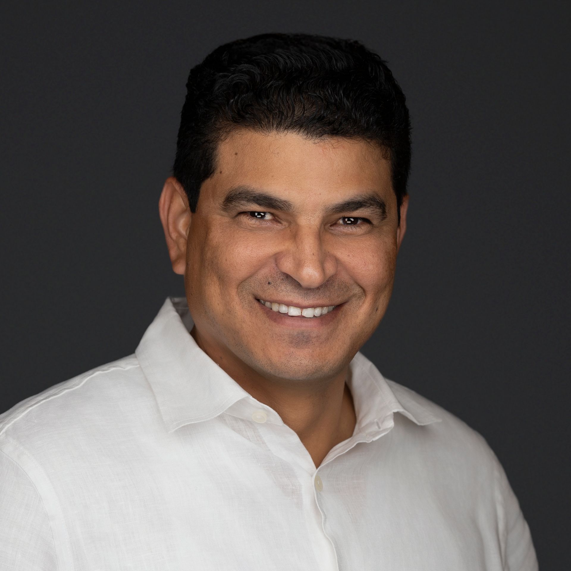 Man smiling, wearing a white shirt, against a gray backdrop.