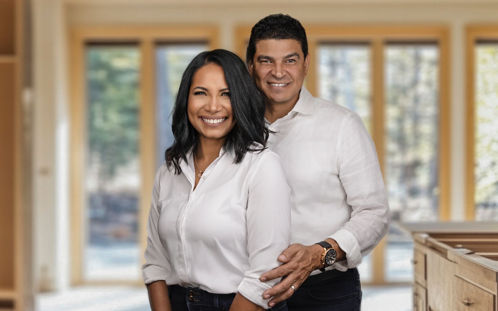 Couple smiling, posing in an unfinished room with large windows; man's arm around woman's waist.