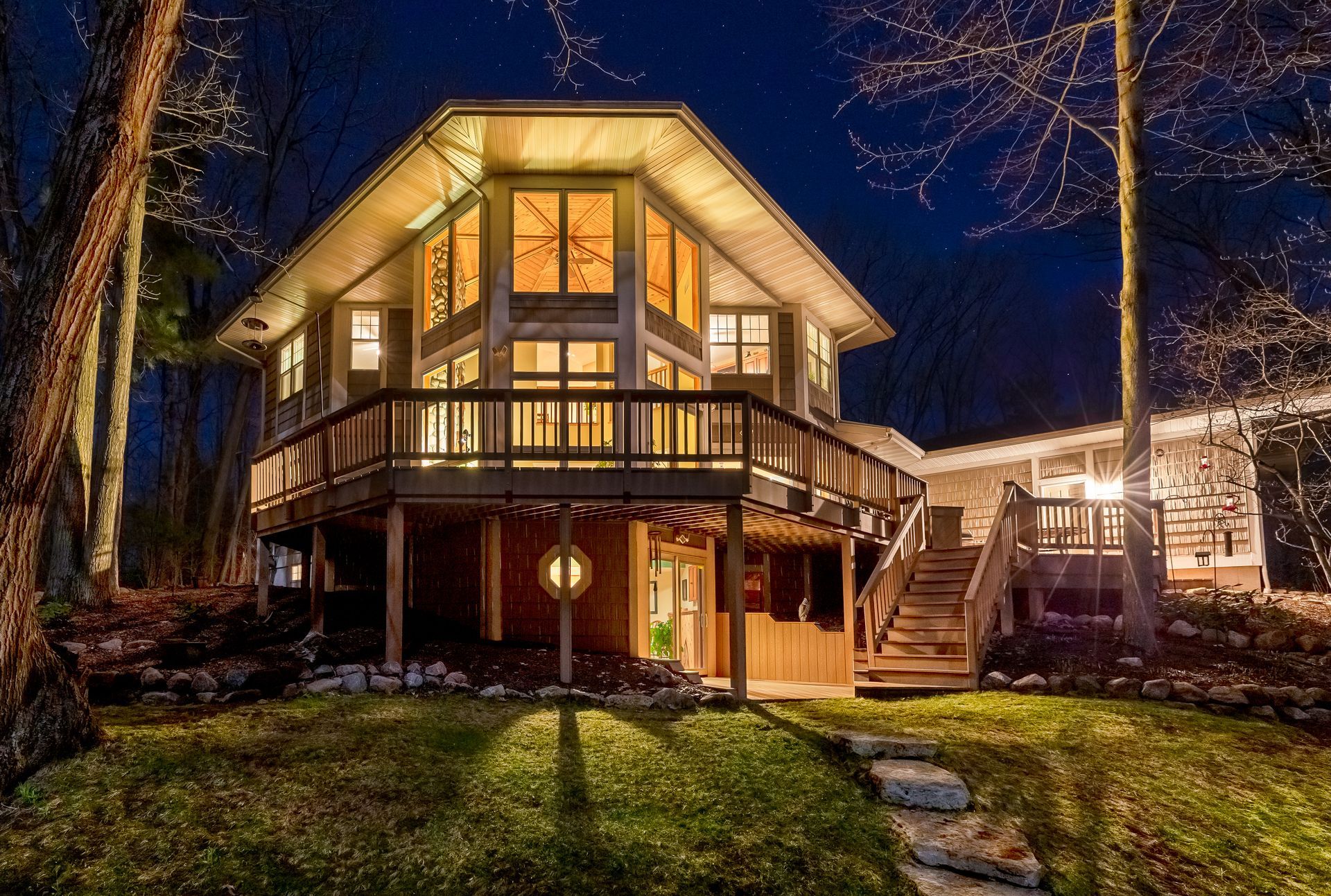 Lit wooden cabin with large windows and wrap-around deck at night.