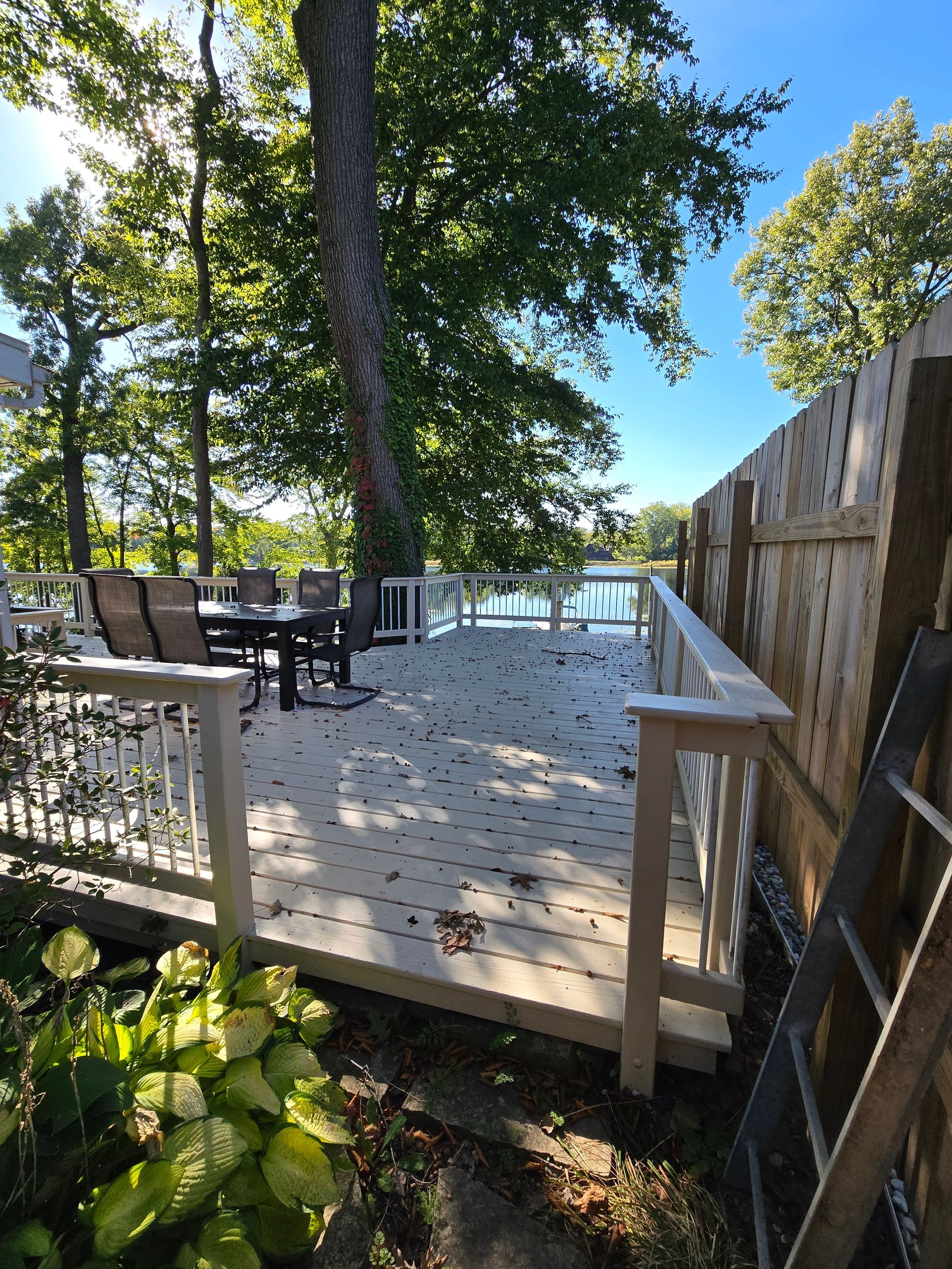 A wooden deck with white railings overlooks a lake, featuring outdoor furniture under large trees on a sunny day.