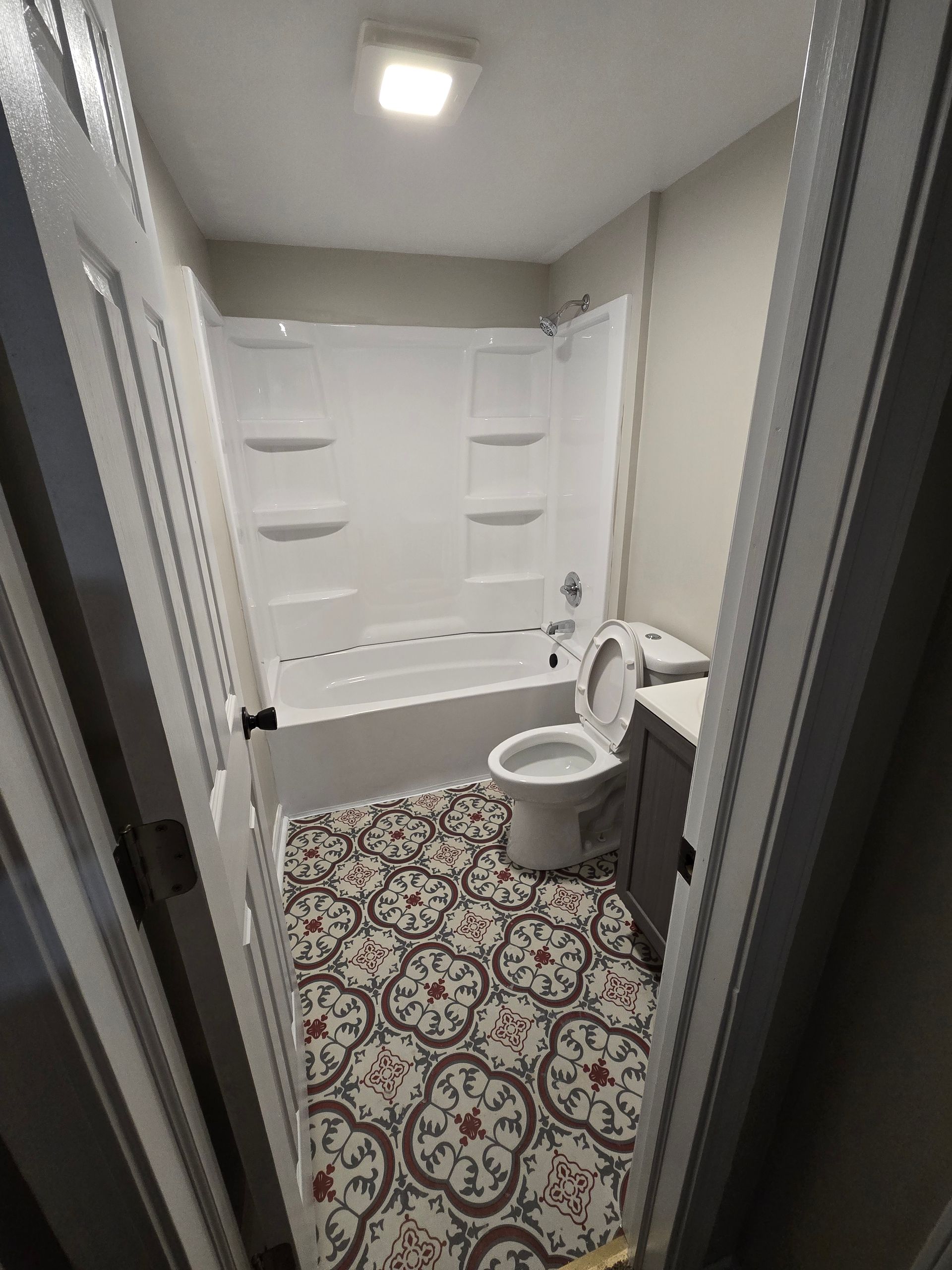 A bathroom viewed through a doorway, featuring white bathtub walls, a white toilet, and patterned tile flooring.