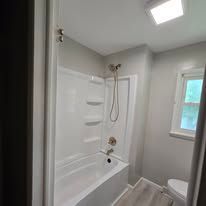 A bathroom featuring a white bathtub and shower surround, light gray walls, a window, and a white toilet.