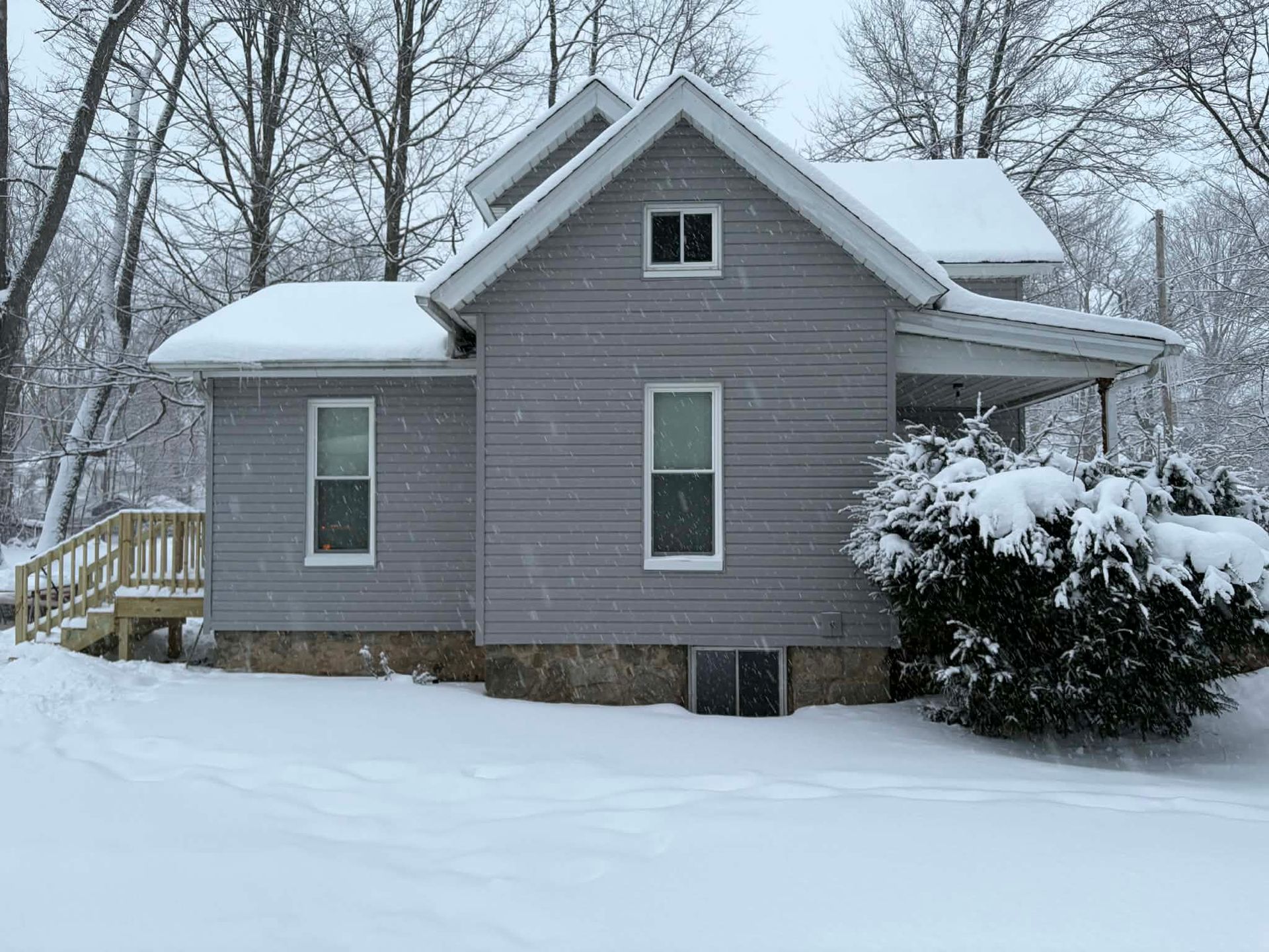 A grey, two-story house with a stone foundation sits in a snowy yard surrounded by snow-covered trees and a small porch.